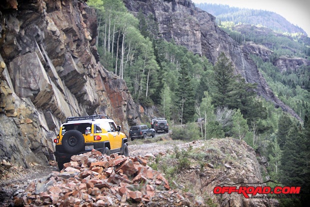Four-Wheel-Drives rolling along jagged rocks and steep cliffs, surrounded by conifers and aspen trees in the San Juan Mountains, Colorado.